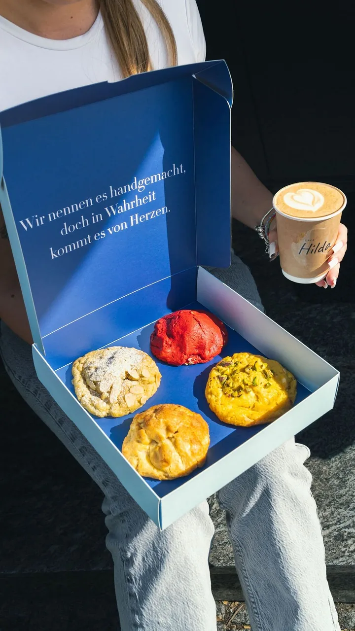 Geöffnete Cookie-Box von Café Hilda auf dem Schoß, daneben ein Cappuccino